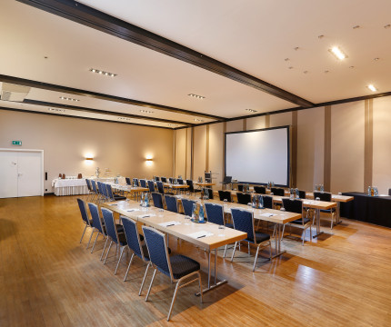 Konferenzraum mit blauen Stühlen, langer Tischreihe und Leinwand an der Wand