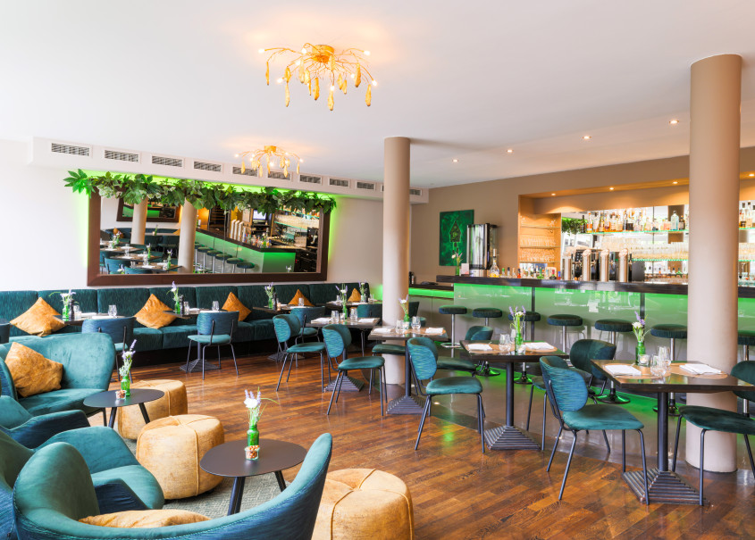 Blick auf den Lounge Bereich, im Hintergrund schön ein gedeckte Tische und die Bar, alles in dunklen Grüntönen sehr modern